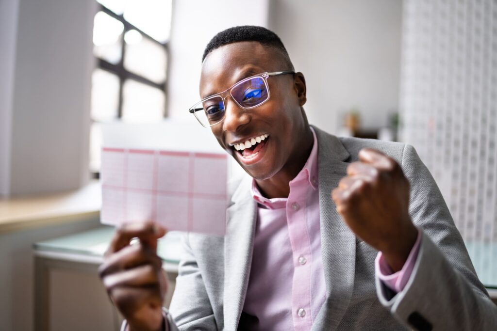 Ein glücklicher Mann im Anzug hält einen Lottoschein in der Hand und feiert seinen Gewinn beim Lotto online spielen in einem hellen Büro.