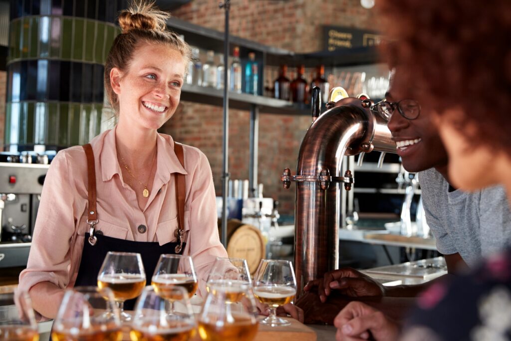 Eine Gruppe von Freunden genießt ein Biertasting in einer gemütlichen Bar, geführt von einer lächelnden Barkeeperin mit einer Auswahl an verschiedenen Bieren in Verkostungsgläsern.