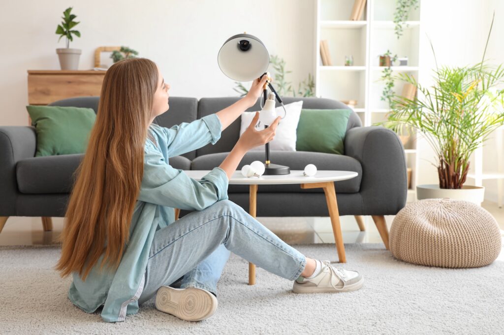 Junge Frau sitzt auf dem Boden eines gemütlichen Wohnzimmers und wechselt die Glühbirne einer Tischlampe.