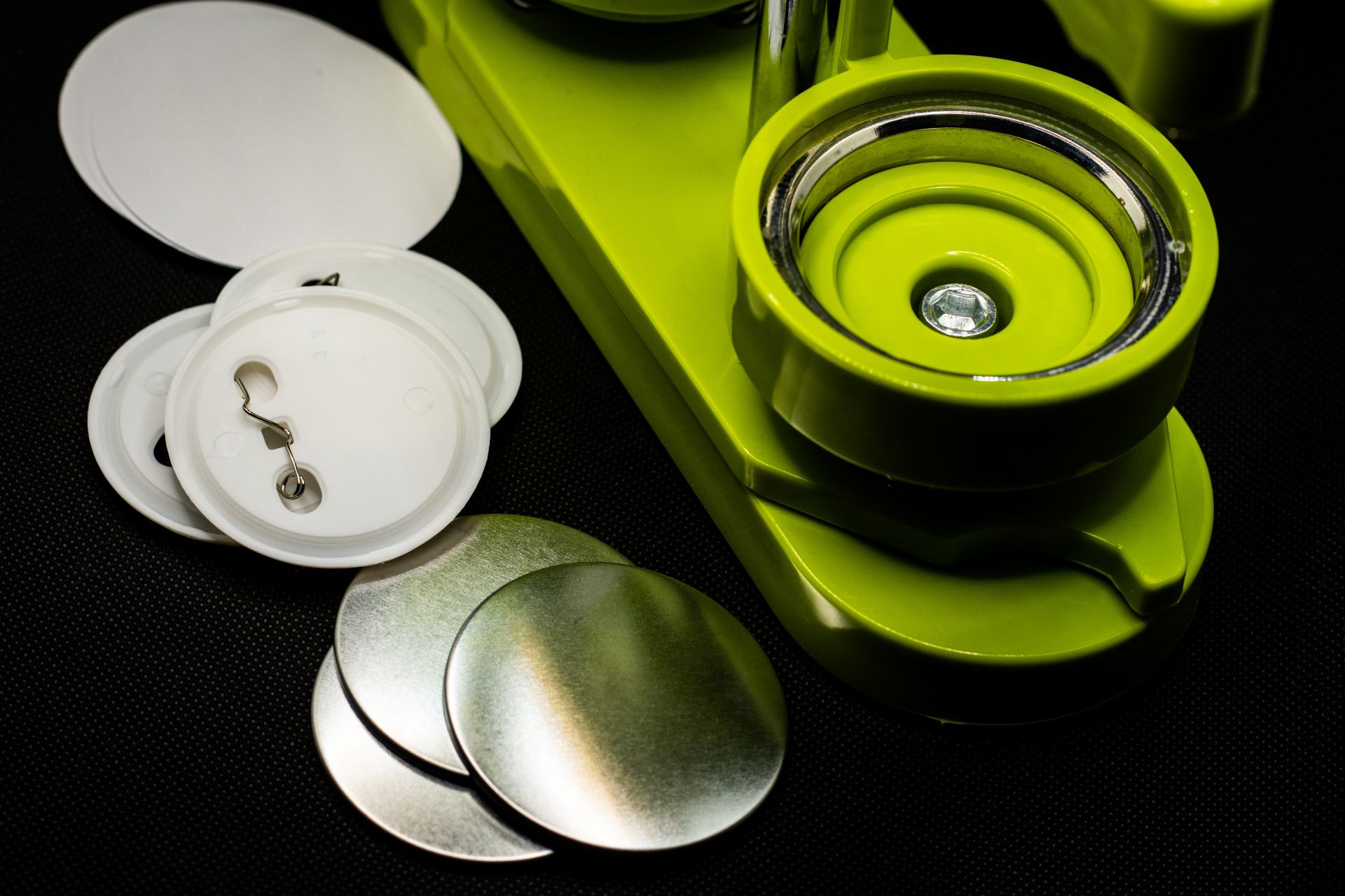Nahaufnahme einer grünen Buttonmaschine mit Rohlingen, Metallplatten und Nadelverschluss – Fokus auf Technik und Zubehör.