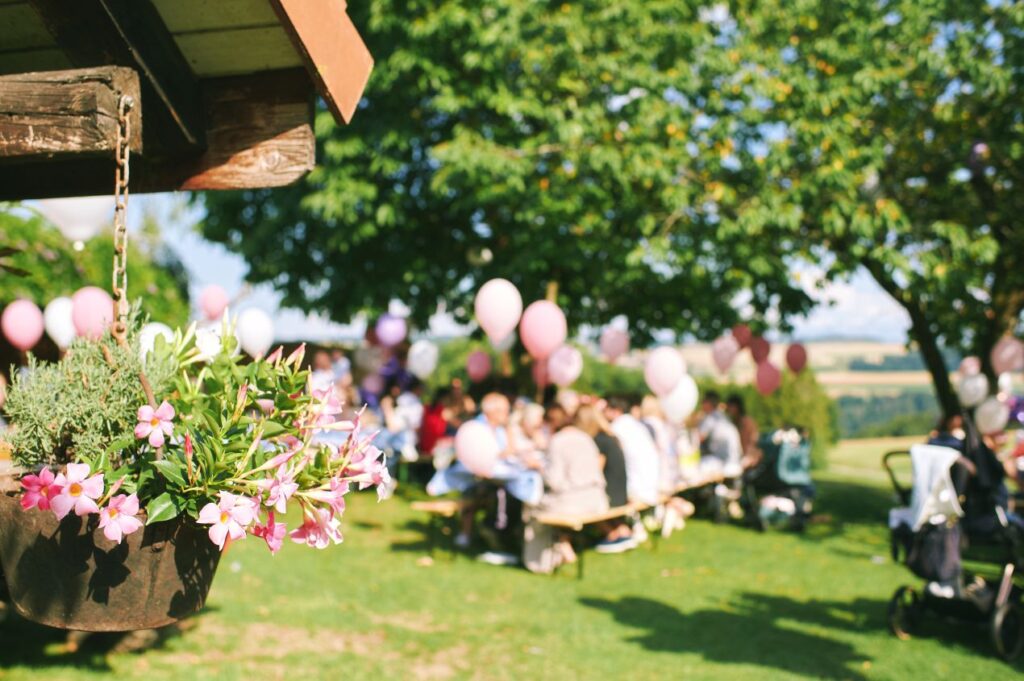 Helles Foto einer Bauernhof Feier mit Blumen im Vordergrund, Gaesten an langen Tischen und rosa Ballons unter schattigen Baeumen